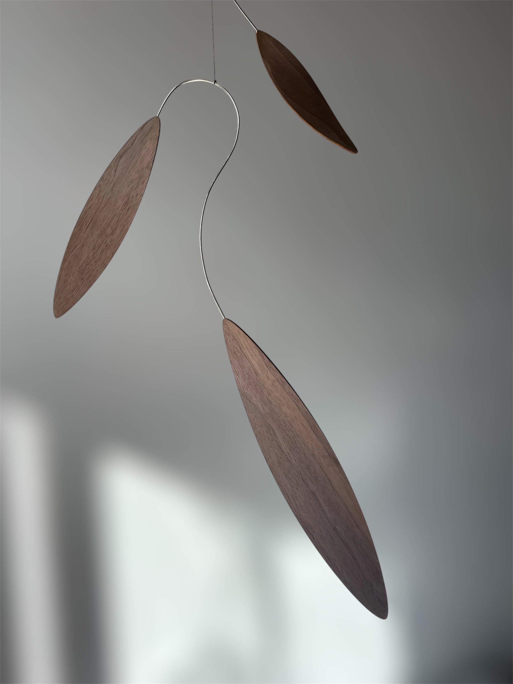 Large 1 metre walnut leaf mobile hanging on a wall beside artist, handcrafted layered walnut veneer wooden mobile with curved leaf forms and stainless steel wire, modern kinetic sculpture, moving art for adults, mid century decor, Scandinavian design minimalist decor, Calder inspired art mobile, unique designer home decor, wood hanging art for living room, gallery wall decor, contemporary mobile handmade in the UK by The Illuminist, soothing balancing art and modern wood art