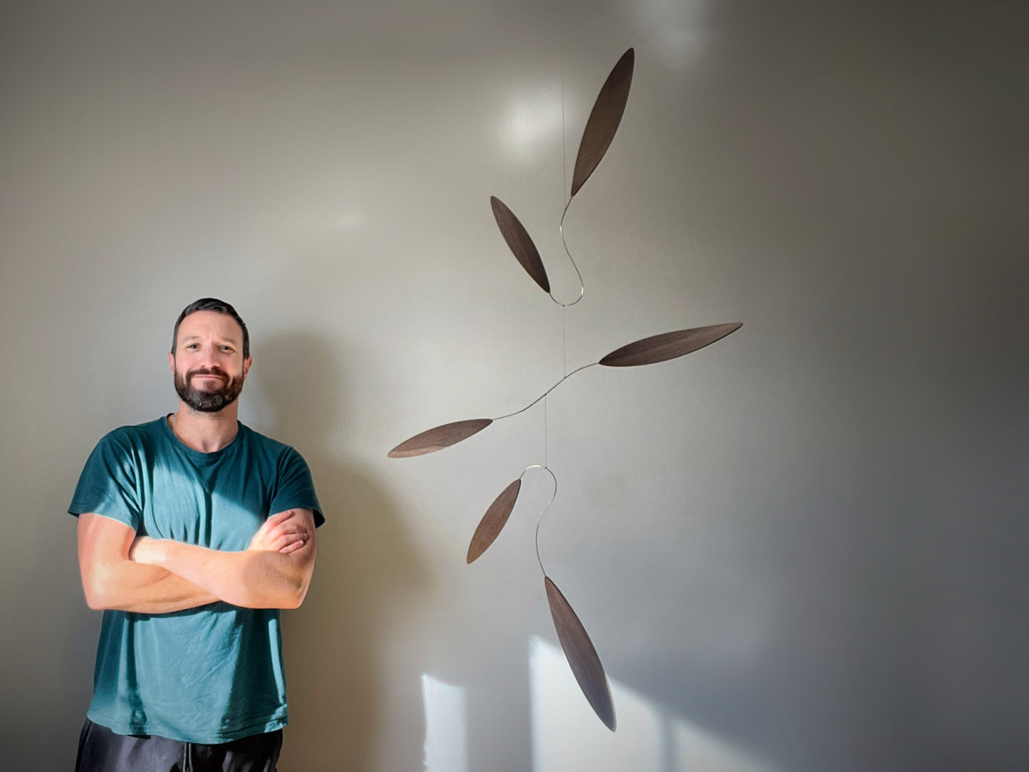 Large 1 metre walnut leaf mobile hanging on a wall beside artist, handcrafted layered walnut veneer wooden mobile with curved leaf forms and stainless steel wire, modern kinetic sculpture, moving art for adults, mid century decor, Scandinavian design minimalist decor, Calder inspired art mobile, unique designer home decor, wood hanging art for living room, gallery wall decor, contemporary mobile handmade in the UK by The Illuminist, soothing balancing art and modern wood art