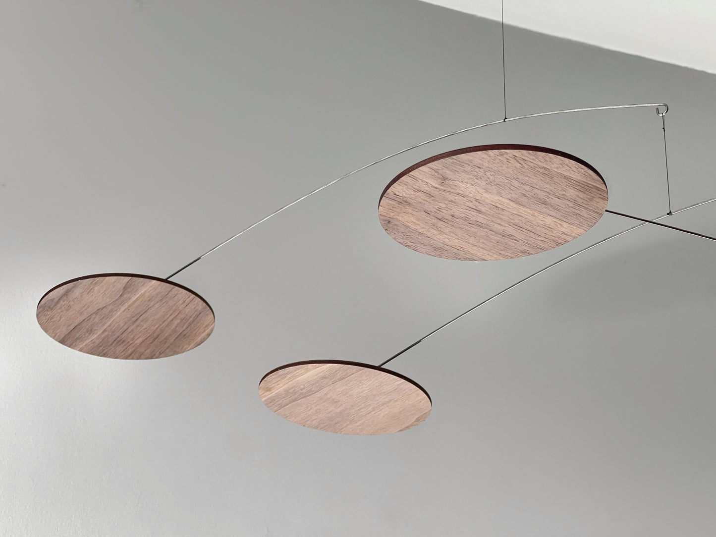 modern ceiling sculpture, kinetic mobile features multiple wooden discs in dark walnut finish, suspended by thin metal wire and balanced arms. The discs cast long shadows against a light grey wall, showcasing a delicate balance and mid-century modern or minimalist design. The assembly is an Alexander Calder-style mobile, emphasizing movement and negative space.