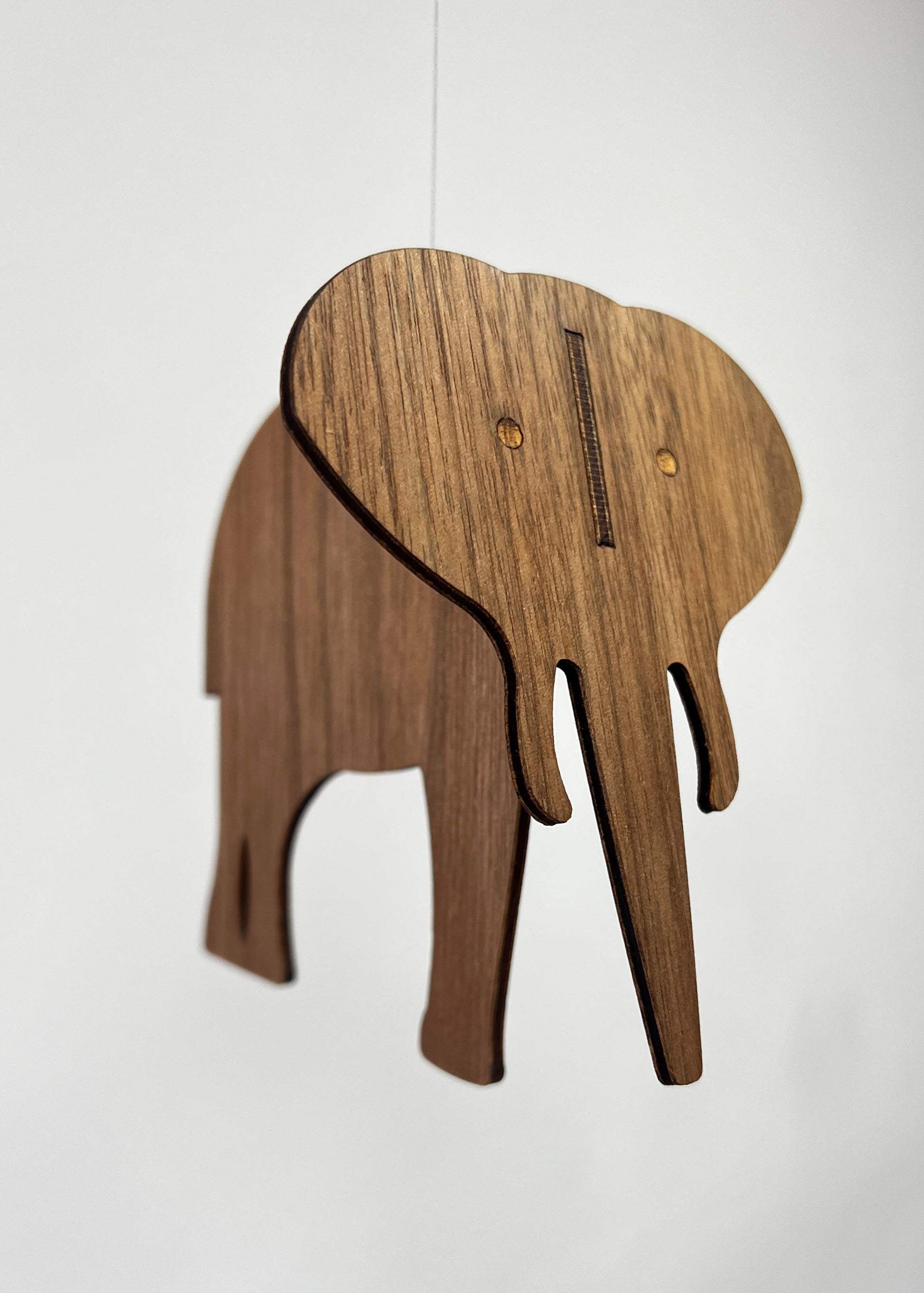 Close-up of the handmade elephant mobile piece cut from engineered walnut wood showing detailed wood grain and simple modern silhouette. The elephant forms part of an African animal mobile designed for nursery decor and baby mobile use above a crib or cot. Suspended with fine thread as part of a balanced hanging mobile inspired by Scandinavian Flensted mobile design. This wooden elephant mobile element pairs with a giraffe mobile and lion mobile to create a safari themed Africa mobile.