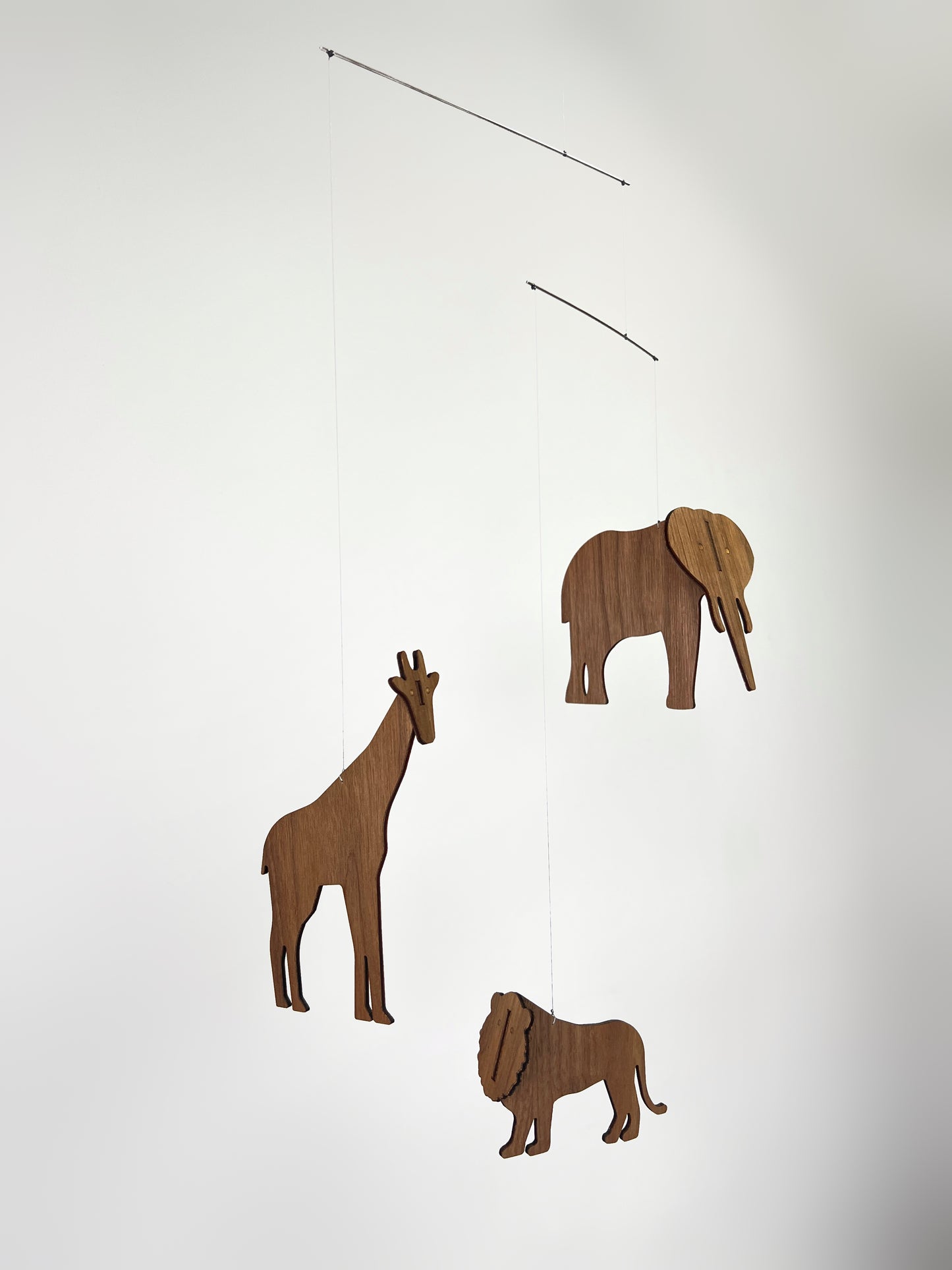African animal baby mobile hanging above a white cot creating soothing movement for a newborn nursery. This handmade wooden mobile features an elephant, giraffe and lion cut from engineered walnut wood, suspended as a balanced hanging mobile. Ideal nursery decor for safari or Africa themed rooms, the animal mobile gently rotates with air movement similar to a classic Flensted mobile. Designed for use as a cot mobile or crib decoration, it makes a beautiful baby gift and statement nursery mobile