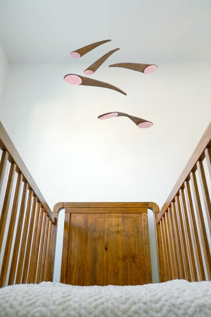 in blush pink this alternative baby mobile in rich walnut wood, featuring curved forms and a floating kinetic structure. This handcrafted nursery decor piece rotates gently, creating a peaceful sensory experience for babies. Inspired by vintage mid century modern design, it works in minimalist and design-led homes. A unique heirloom gift with custom colours available.