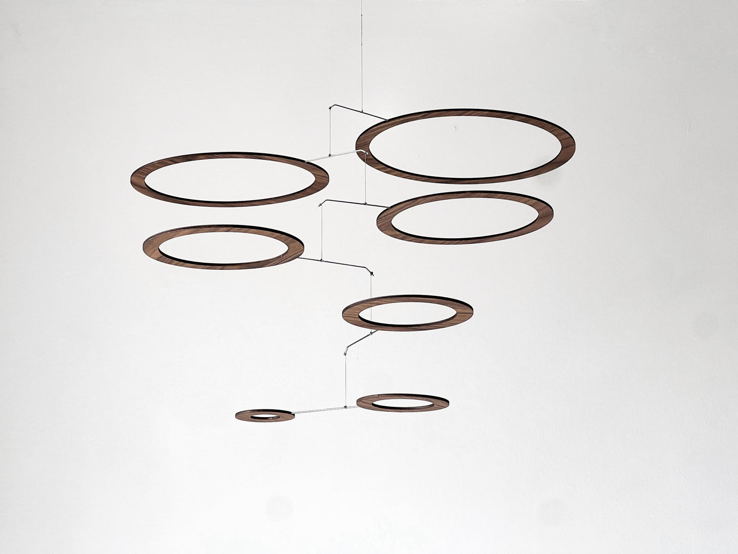 Handmade walnut wood circle mobile hanging above a minimalist home office desk. Seven layered walnut veneer rings on stainless steel rods catch natural light from a nearby window. A calm, sculptural ceiling feature for modern home offices, Scandinavian interiors, and design-led spaces. Approx 67cm wide × 30cm tall (26.5 × 11.8 in).