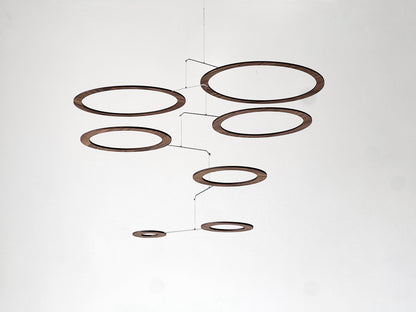 Handmade walnut wood circle mobile hanging above a minimalist home office desk. Seven layered walnut veneer rings on stainless steel rods catch natural light from a nearby window. A calm, sculptural ceiling feature for modern home offices, Scandinavian interiors, and design-led spaces. Approx 67cm wide × 30cm tall (26.5 × 11.8 in).