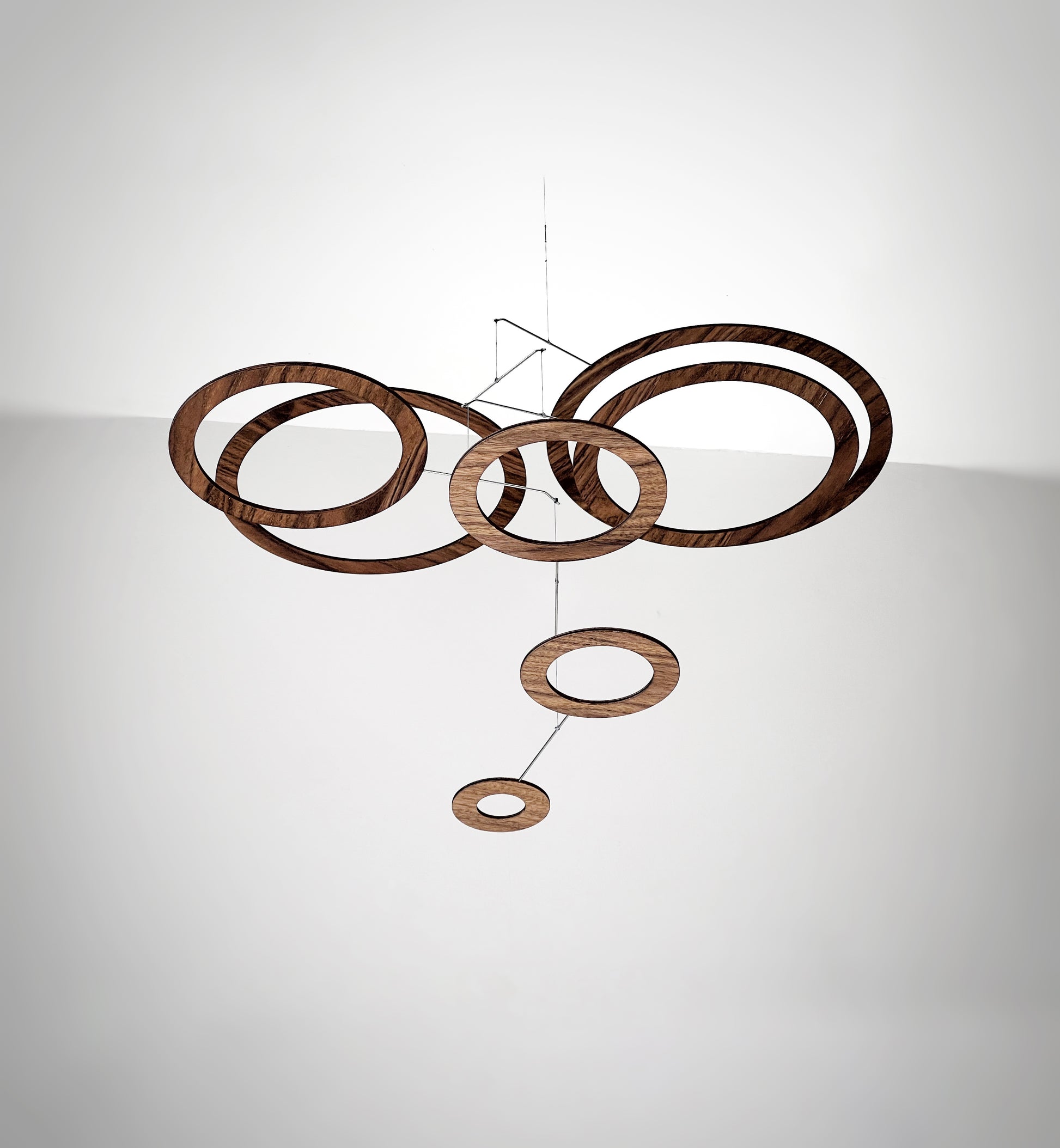 Handmade walnut wood circle mobile hanging above a minimalist home office desk. Seven layered walnut veneer rings on stainless steel rods catch natural light from a nearby window. A calm, sculptural ceiling feature for modern home offices, Scandinavian interiors, and design-led spaces. Approx 67cm wide × 30cm tall (26.5 × 11.8 in).