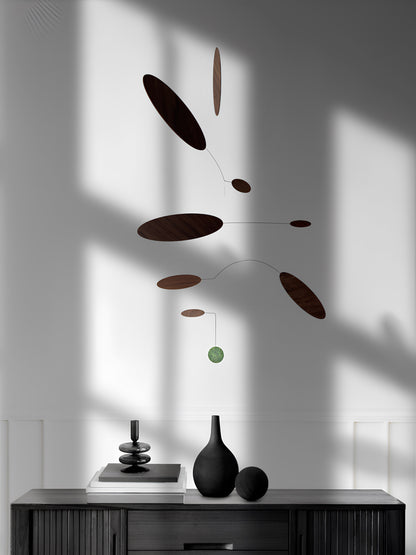 A modern mobile wall hanging with dark brown oval discs and a small green sphere suspended from thin wires, displayed against a white wall with dramatic window shadows. Below, a dark console table holds minimalist black vases and stacked books.