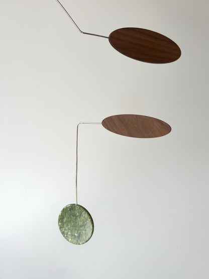 A minimalist mobile hanging against a plain light background, featuring two dark brown walnut wooden oval discs suspended by thin metal rods from a textured green jade stone circular disc.