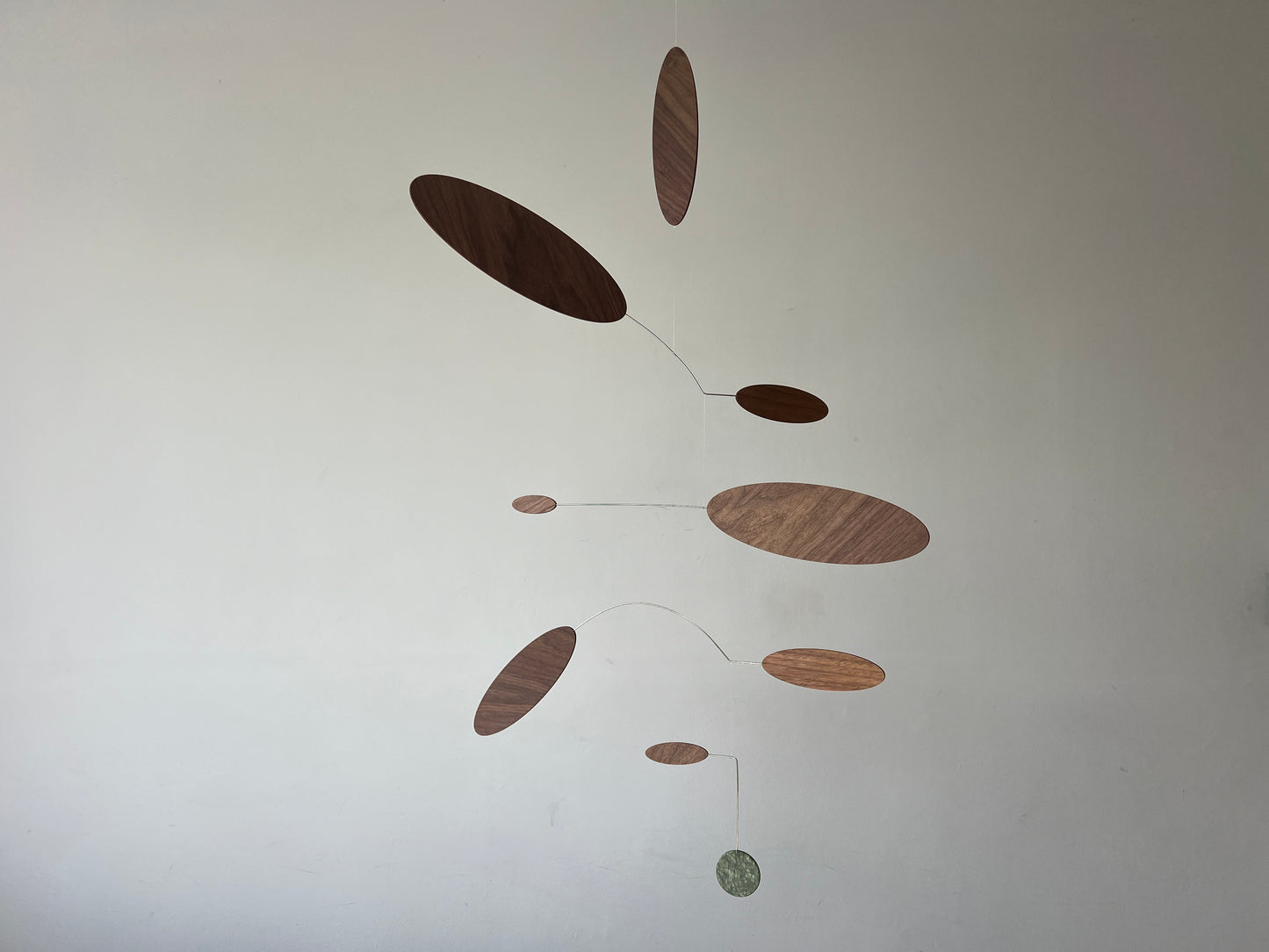 A minimalist mobile wall hanging with multiple oval and circular wooden discs in varying wood tones, suspended by thin wires against a plain off-white wall.