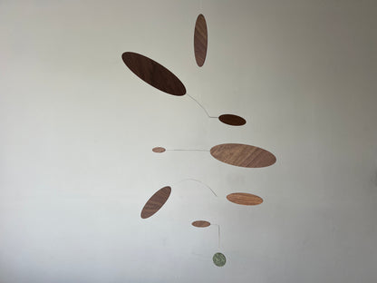 A minimalist mobile wall hanging with multiple oval and circular wooden discs in varying wood tones, suspended by thin wires against a plain off-white wall.