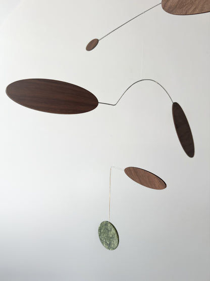 A minimalist mobile with organic-shaped wooden and stone discs suspended on thin metal wires against a plain white background.