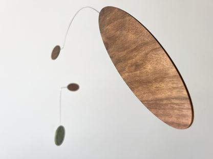 A minimalist mobile wall hanging with a large oval wooden disc on a curved wire, accompanied by two smaller hanging elements, photographed against a plain light background.