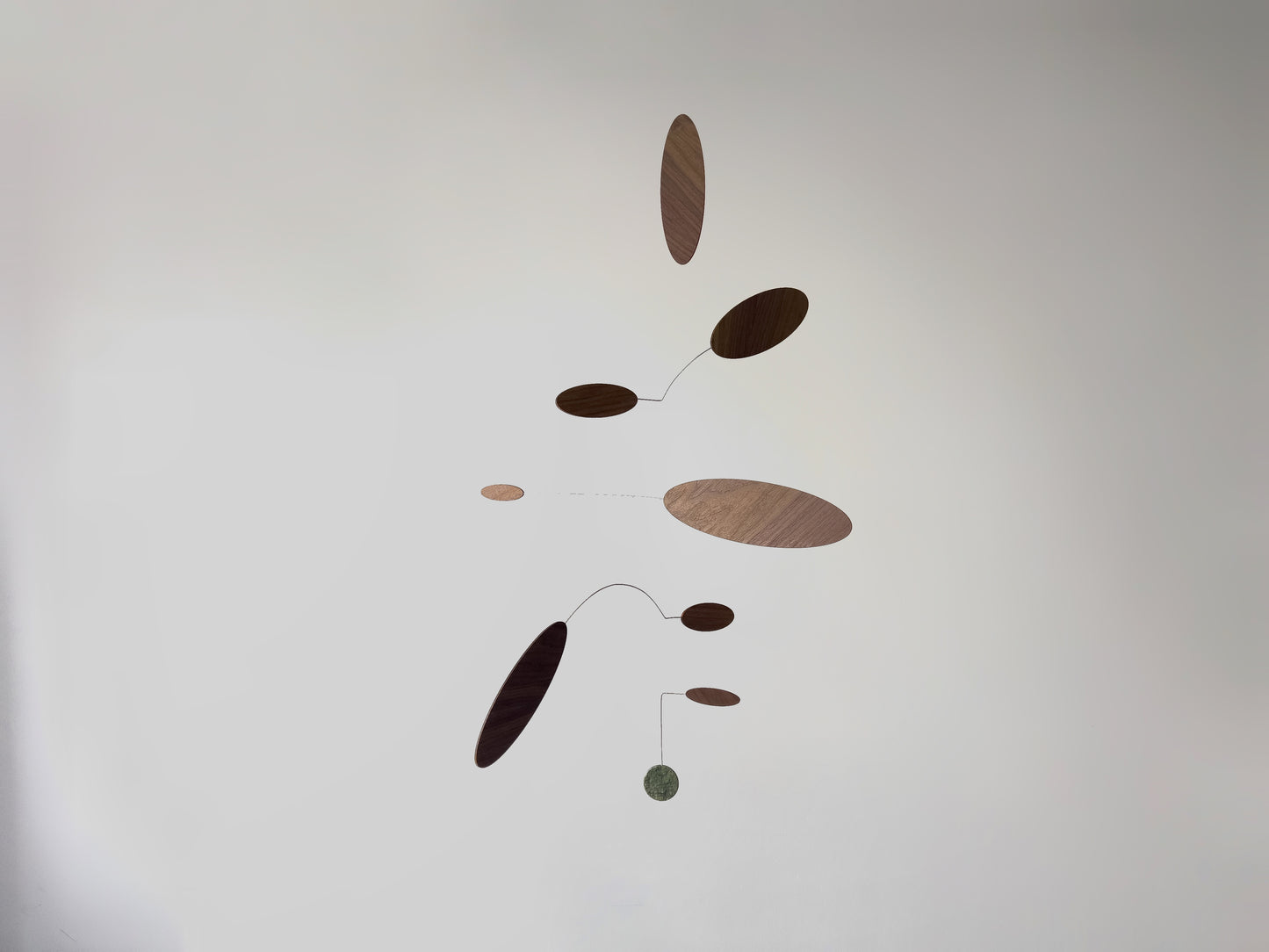 A minimalist mobile with floating oval and circular wooden weights on thin curved wires, suspended against a plain light gray wall.