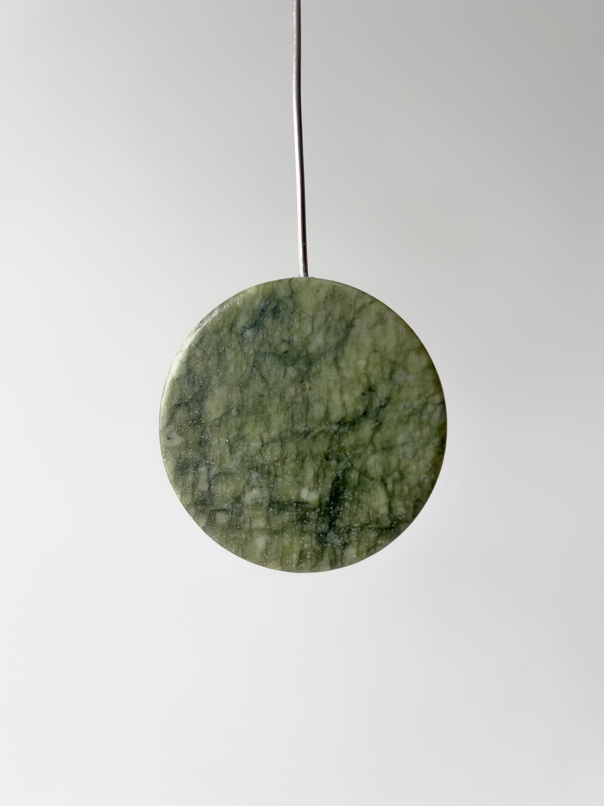A round green stone pendant with natural marbled texture, suspended by a thin black cord against a light gray background.