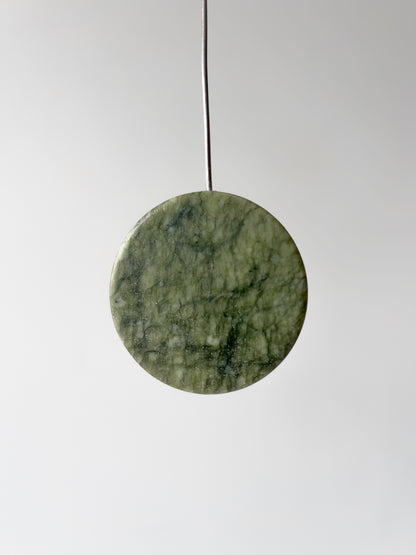 A round green stone pendant with natural marbled texture, suspended by a thin black cord against a light gray background.