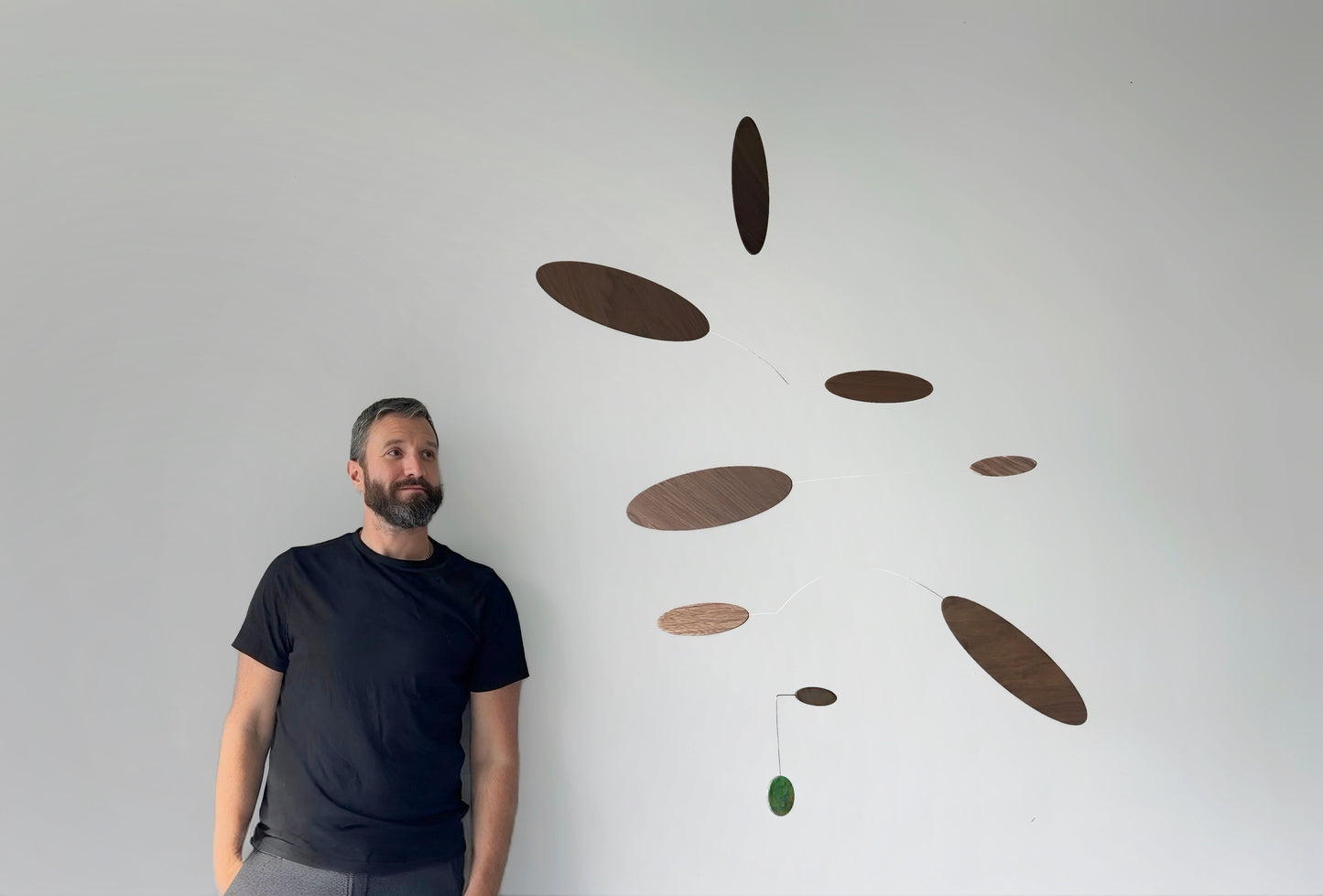 A man wearing a black t-shirt and gray pants stands next to a minimalist mobile wall art piece made of floating wooden oval discs in various shades of brown, with a small green jade stone hanging accent, against a plain white wall.