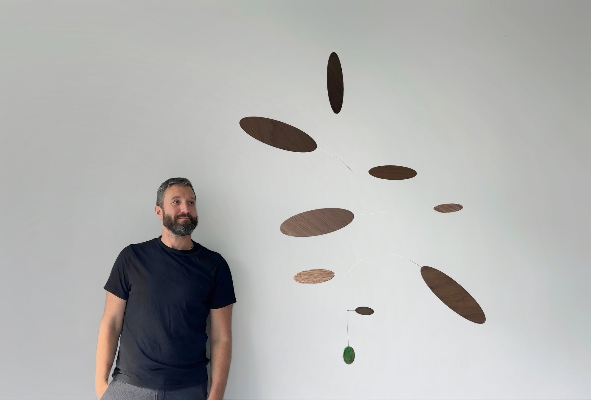 A man wearing a black t-shirt and gray pants stands next to a minimalist mobile wall art piece made of floating wooden oval discs in various shades of brown, with a small green jade stone hanging accent, against a plain white wall.
