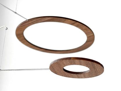 Handmade walnut wood circle mobile hanging above a minimalist home office desk. Five layered walnut veneer rings on stainless steel rods catch natural light from a nearby window. A calm, sculptural ceiling feature for modern home offices, Scandinavian interiors, and design-led spaces. Approx 67cm wide × 30cm tall (26.5 × 11.8 in).