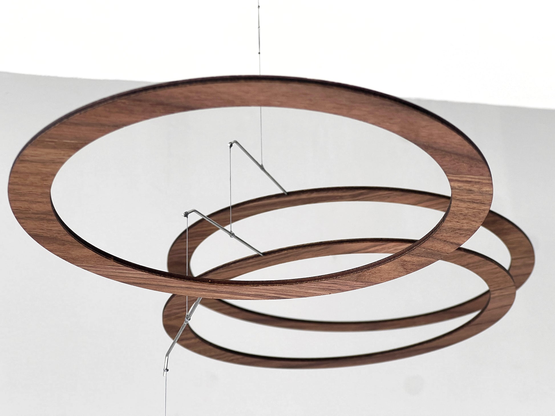 Handmade walnut wood circle mobile hanging above a minimalist home office desk. Seven layered walnut veneer rings on stainless steel rods catch natural light from a nearby window. A calm, sculptural ceiling feature for modern home offices, Scandinavian interiors, and design-led spaces. Approx 67cm wide × 30cm tall (26.5 × 11.8 in).
