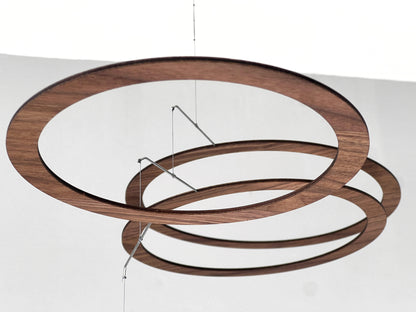 Handmade walnut wood circle mobile hanging above a minimalist home office desk. Seven layered walnut veneer rings on stainless steel rods catch natural light from a nearby window. A calm, sculptural ceiling feature for modern home offices, Scandinavian interiors, and design-led spaces. Approx 67cm wide × 30cm tall (26.5 × 11.8 in).