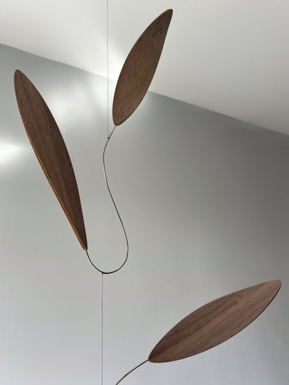 Large 1 metre walnut leaf mobile hanging on a wall beside artist, handcrafted layered walnut veneer wooden mobile with curved leaf forms and stainless steel wire, modern kinetic sculpture, moving art for adults, mid century decor, Scandinavian design minimalist decor, Calder inspired art mobile, unique designer home decor, wood hanging art for living room, gallery wall decor, contemporary mobile handmade in the UK by The Illuminist, soothing balancing art and modern wood art