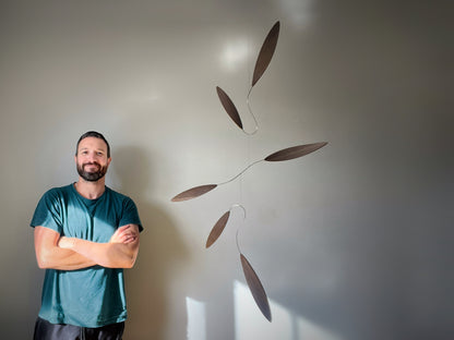 Large 1 metre walnut leaf mobile hanging on a wall beside artist, handcrafted layered walnut veneer wooden mobile with curved leaf forms and stainless steel wire, modern kinetic sculpture, moving art for adults, mid century decor, Scandinavian design minimalist decor, Calder inspired art mobile, unique designer home decor, wood hanging art for living room, gallery wall decor, contemporary mobile handmade in the UK by The Illuminist, soothing balancing art and modern wood art