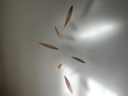 Large 1 metre walnut leaf mobile hanging on a wall beside artist, handcrafted layered walnut veneer wooden mobile with curved leaf forms and stainless steel wire, modern kinetic sculpture, moving art for adults, mid century decor, Scandinavian design minimalist decor, Calder inspired art mobile, unique designer home decor, wood hanging art for living room, gallery wall decor, contemporary mobile handmade in the UK by The Illuminist, soothing balancing art and modern wood art