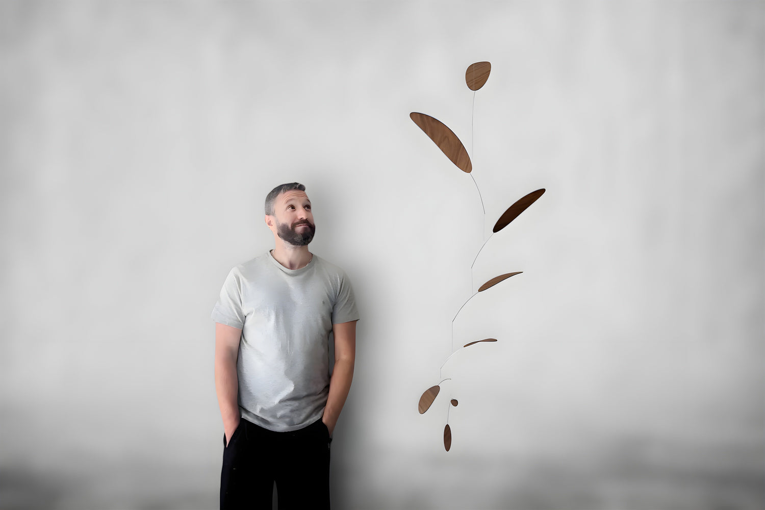 A man with a beard wearing a light gray t-shirt and black pants stands against a textured gray wall, looking up at a floating decorative mobile made of brown leaf-shaped metal pieces.