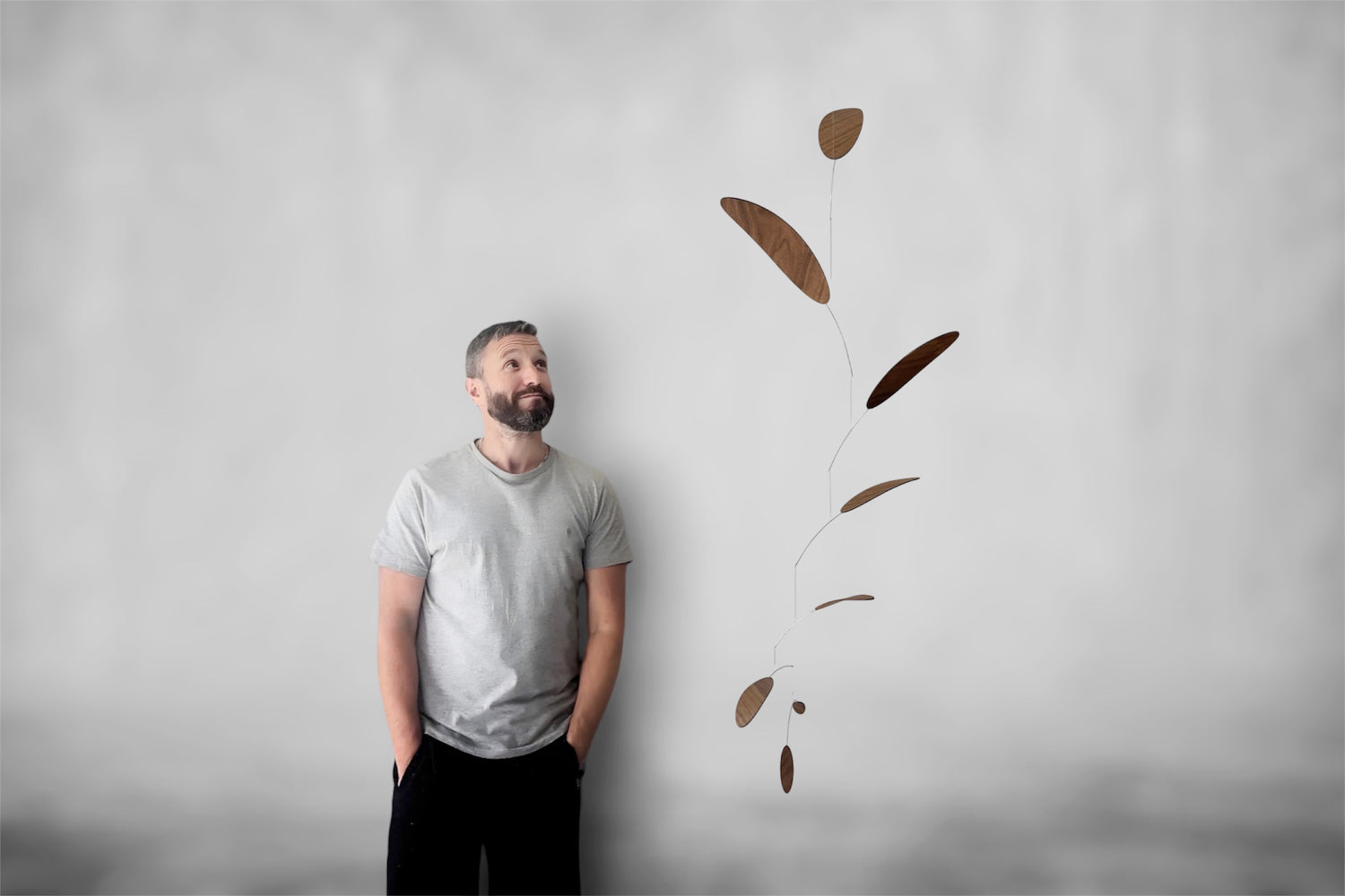 A man in a light gray t-shirt and black pants stands against a textured light gray wall, looking up at a floating mobile made of elongated brown leaf-like shapes suspended by thin wires.