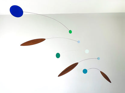 A minimalist mobile with floating wooden teardrop shapes and small colored spheres suspended on thin wires against a plain light gray wall.