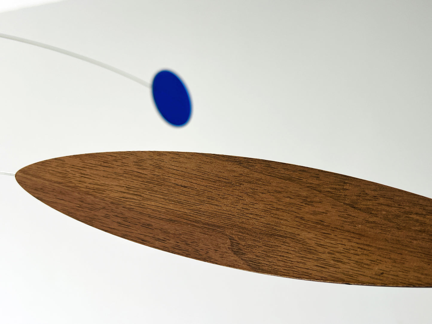 A close-up of a modern mobile or hanging decor piece featuring a large oval wooden wing in warm brown wood grain and a small blue circular weight suspended by a thin white cord against a white background.