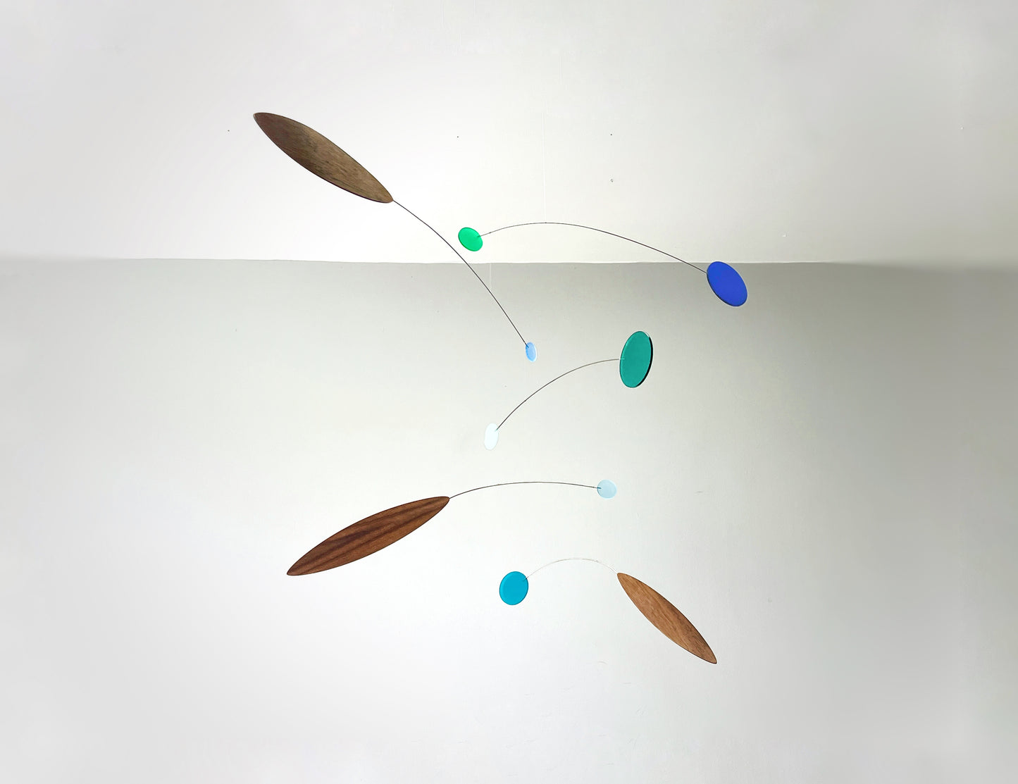 A minimalist calder mobile with floating oval and elongated leaf-like shapes in brown, teal, blue, and light blue, suspended against a plain white wall.