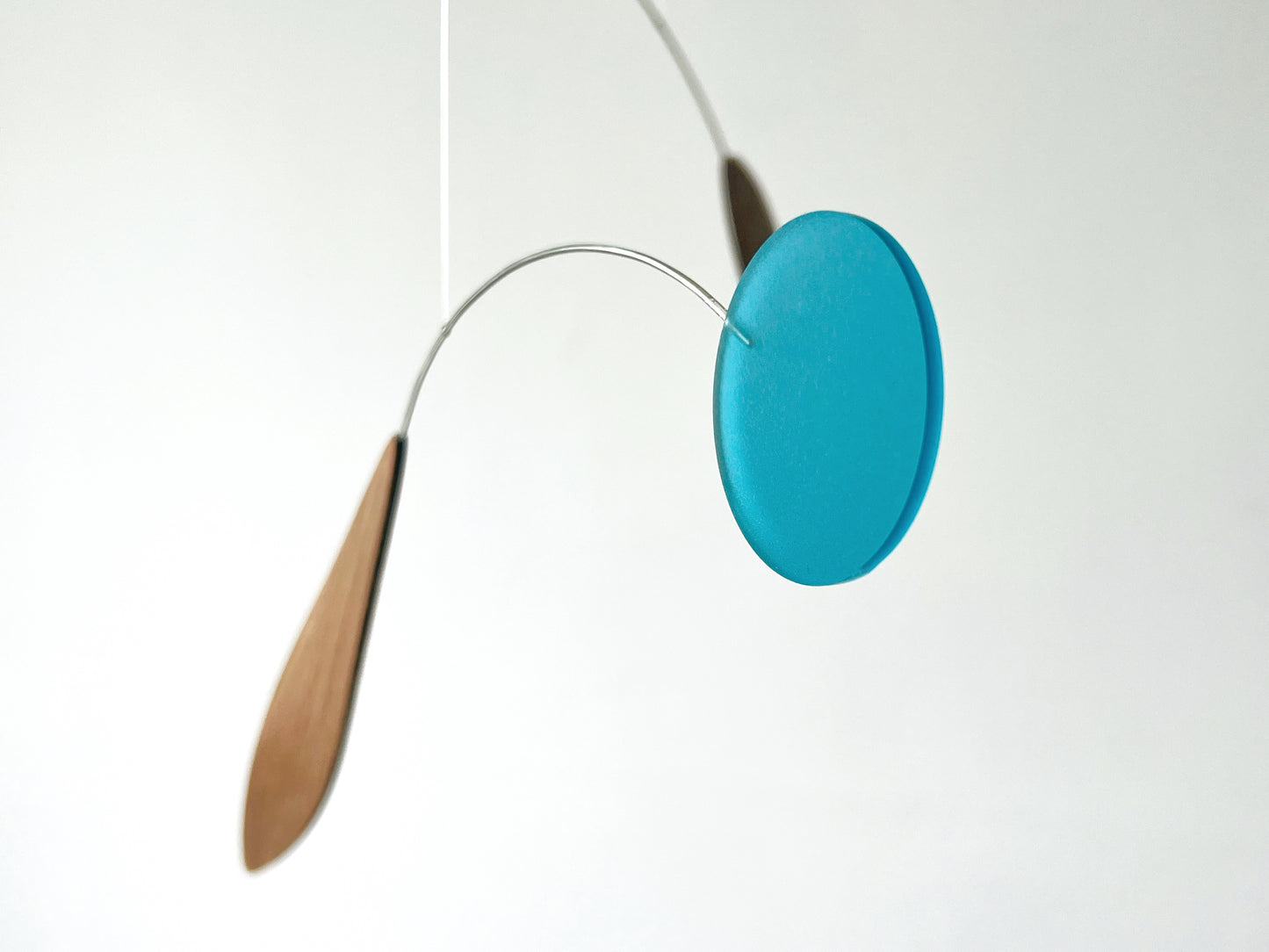 A minimalist mobile hanging from a thin wire, featuring a round translucent turquoise blue disc and two teardrop-shaped wooden weights in light brown, set against a plain white background.
