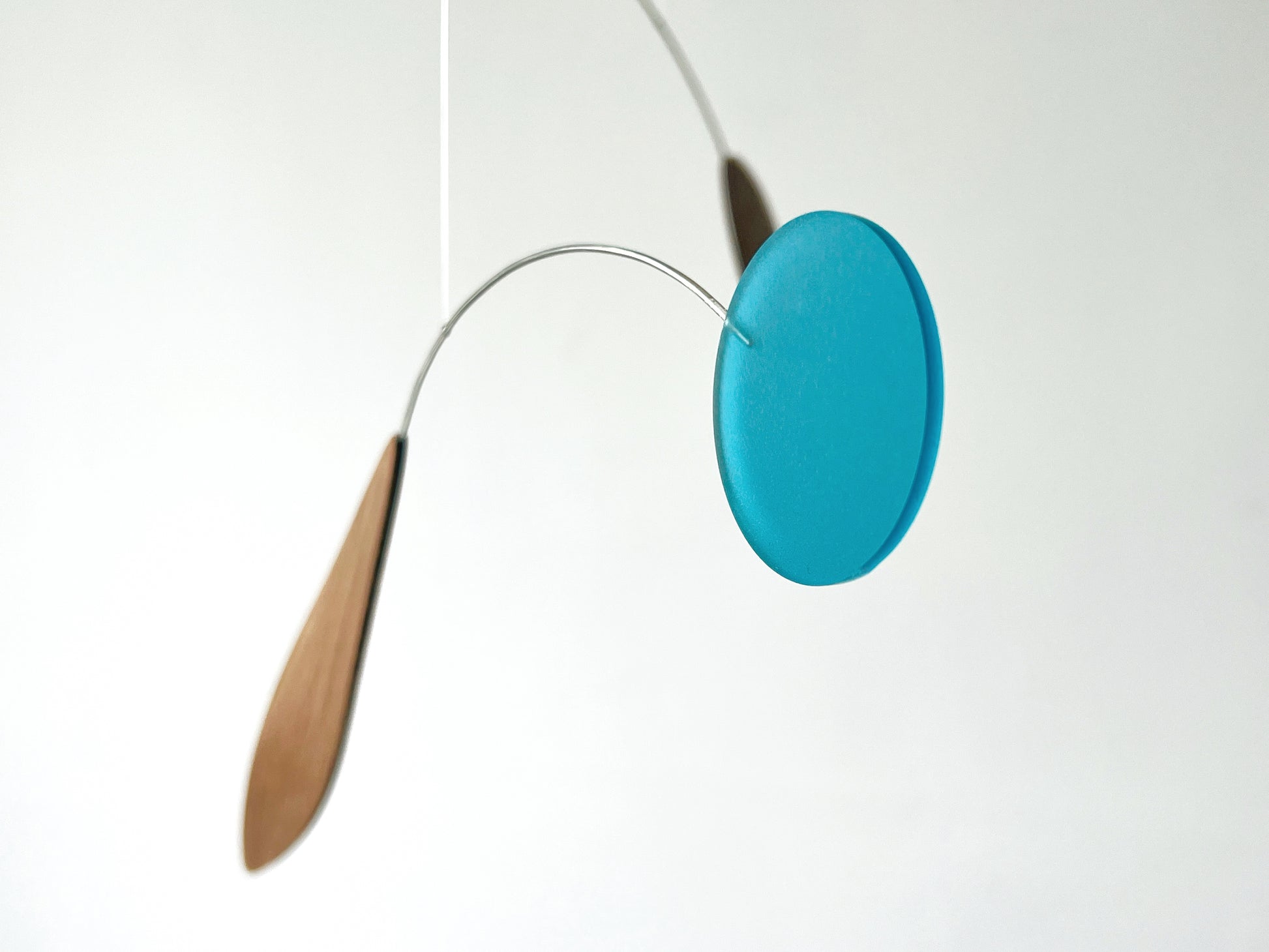 A minimalist mobile hanging from a thin wire, featuring a round translucent turquoise blue disc and two teardrop-shaped wooden weights in light brown, set against a plain white background.