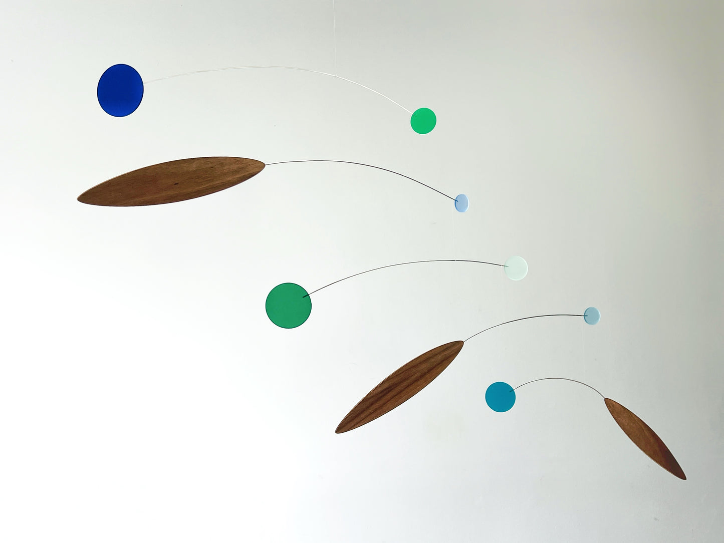 A mid-century modern mobile wall hanging with wooden teardrop-shaped blades and circular discs in blue, green, and light blue, suspended on thin wires against a plain white wall.