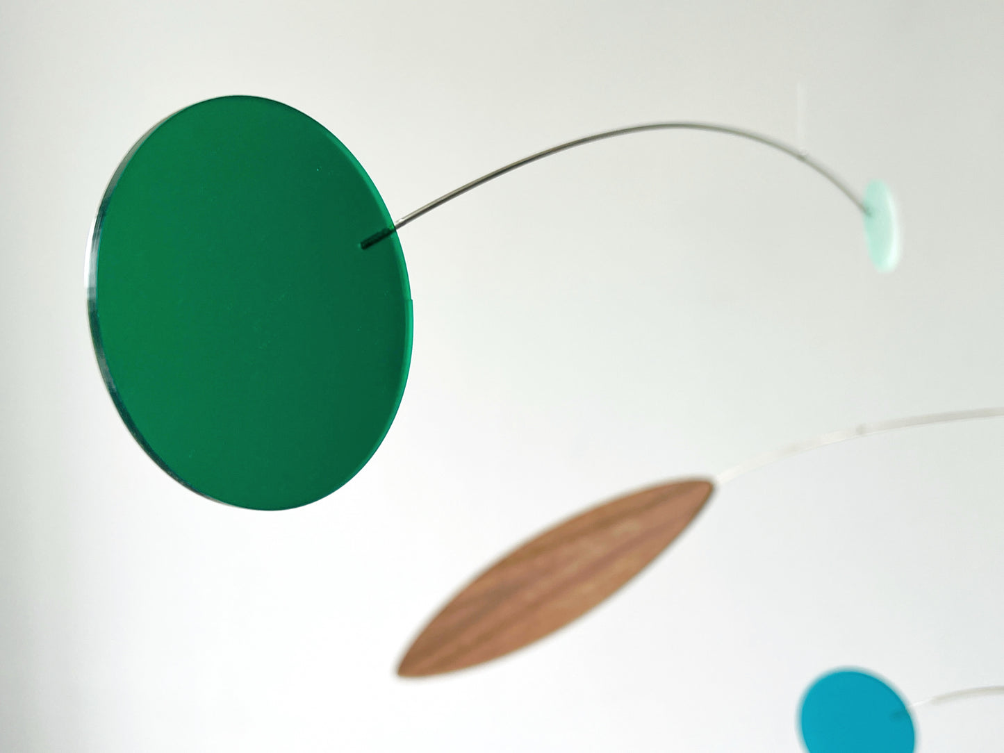 A modern mobile hanging from a thin metal rod, featuring a large green circular disc, a brown oval wood piece, and a small turquoise blue disc against a plain white background.