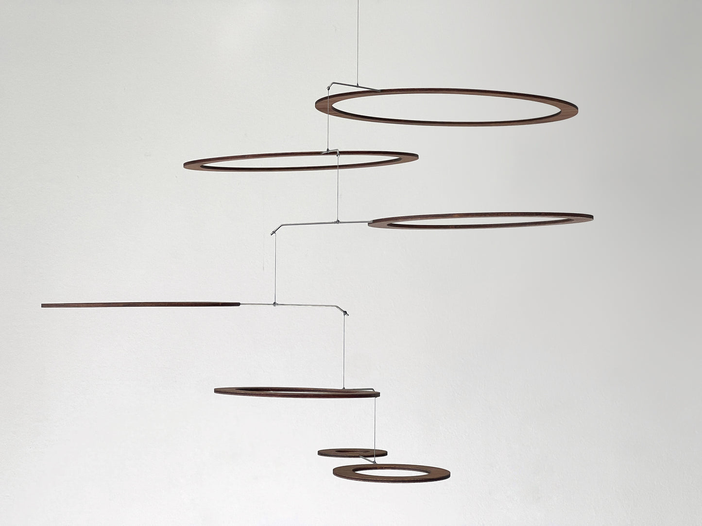 Overhead close-up of seven walnut wood rings in varying sizes, arranged in an interlocking cluster and connected by stainless steel rods and nylon thread. Rich natural grain visible throughout. One ring features a concentric inner circle detail. Handmade kinetic ceiling art ideal for yoga studios, Pilates spaces, and modern Japandi or mid-century interiors.