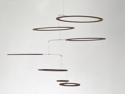 Overhead close-up of seven walnut wood rings in varying sizes, arranged in an interlocking cluster and connected by stainless steel rods and nylon thread. Rich natural grain visible throughout. One ring features a concentric inner circle detail. Handmade kinetic ceiling art ideal for yoga studios, Pilates spaces, and modern Japandi or mid-century interiors.