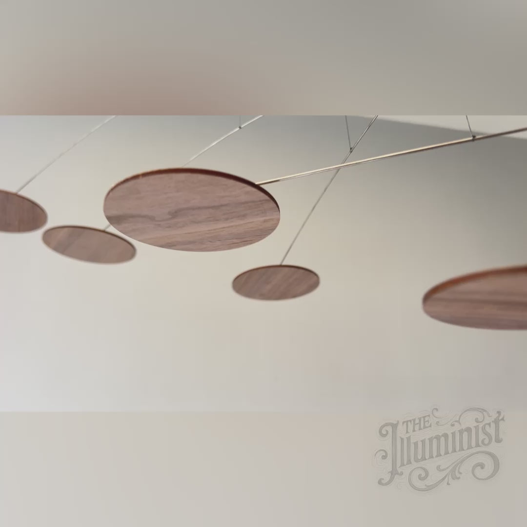 modern ceiling sculpture, kinetic mobile features multiple wooden discs in dark walnut finish, suspended by thin metal wire and balanced arms. The discs cast long shadows against a light grey wall, showcasing a delicate balance and mid-century modern or minimalist design. The assembly is an Alexander Calder-style mobile, emphasizing movement and negative space.